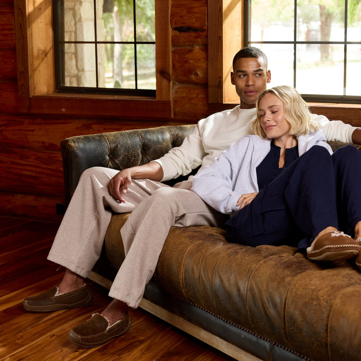 A man and a woman relax in a brown leather tufted sofa inside a rustic log cabin with wood floors and windows. The man wears a cream shirt and beige pants, and the woman wears a light blue cardigan, navy pants, and brown slip-on shoes.