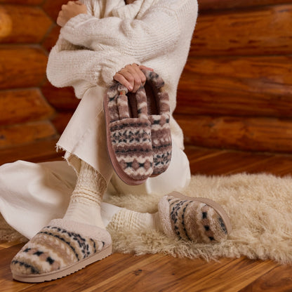A person wrapped in a cream knit sweater sits on a fluffy cream rug in front of a rustic wood log wall, wearing cozy knit socks and slippers with a brown, beige, and gray geometric pattern. They hold one matching slipper in their hand.

