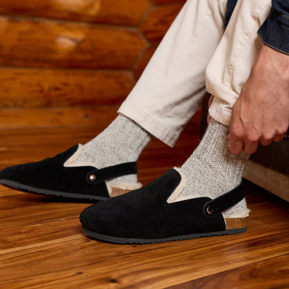 A person sitting in a rustic room wearing black slip-on slippers with elastic ankle straps, light gray knit socks, and cream pants.

