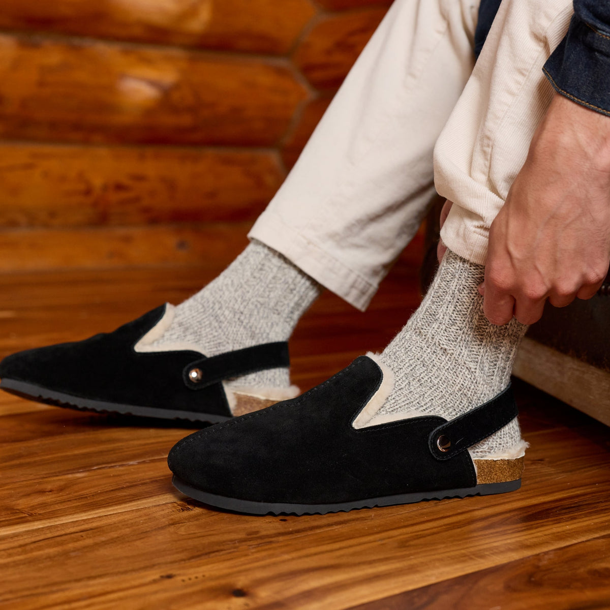 A person sitting in a rustic room wearing black slip-on slippers with elastic ankle straps, light gray knit socks, and cream pants.

