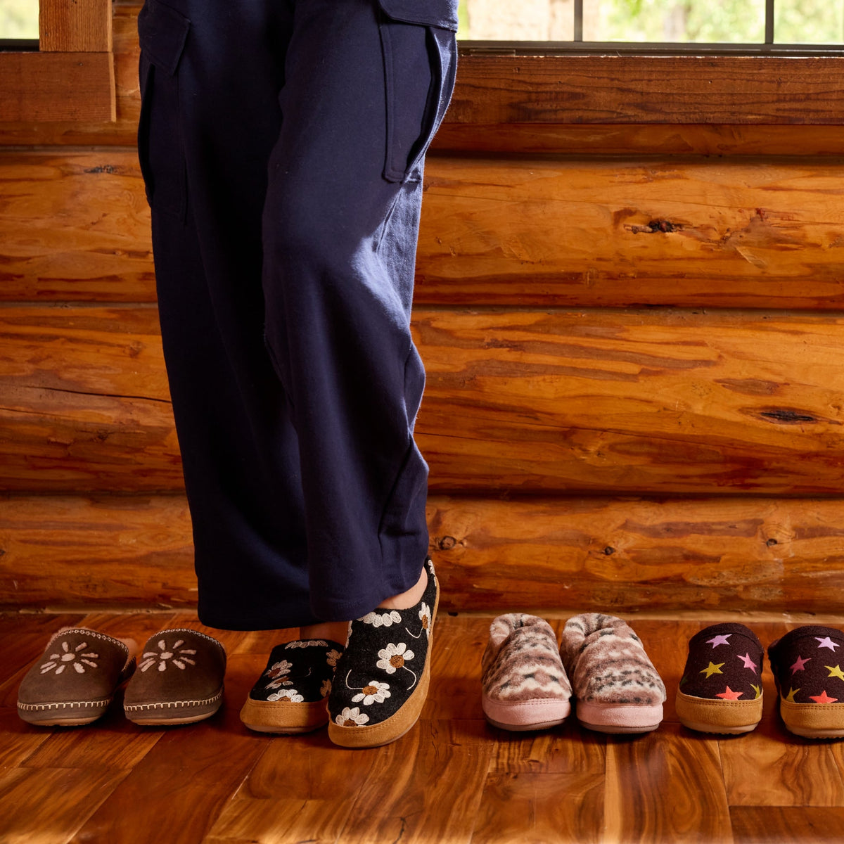 A person wearing dark navy pants stands on a wooden floor in front of a log cabin wall, with six pairs of slip-on mule slippers lined up below. The slippers are in various colors and patterns, including floral, knit, and star motifs.

