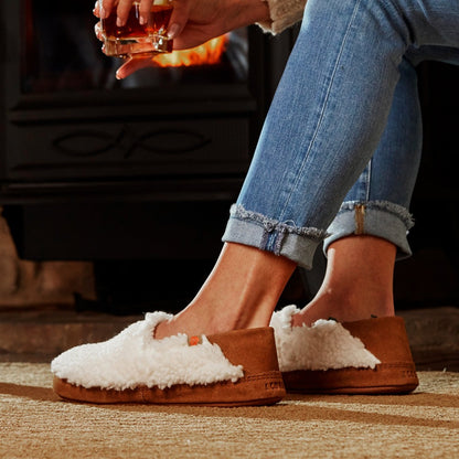 Women’s Acorn Moc with Collapsible Heel Slipper in Buff Popcorn on model sitting by a fireplace with a drink in her hand