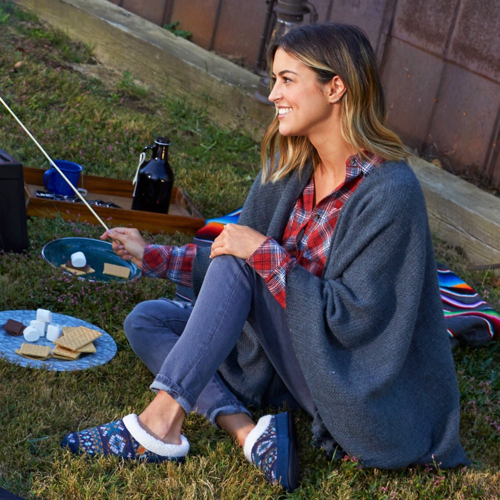 Women’s Fairisles Hoodback Slipper in Blue Multi with different hues of blue and tan on model sitting in front of a barn roasting marshmellows