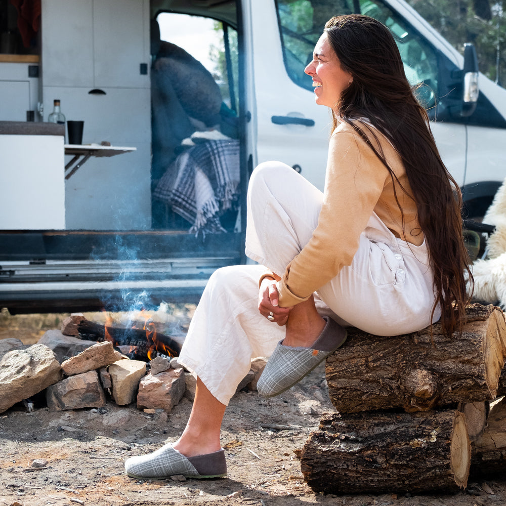 Women’s Algae-Infused Parker Slippers on figure. Model sitting on a pile of wood around a fire. A converted van is behind her. She is laughing and having a good time in her slippers.